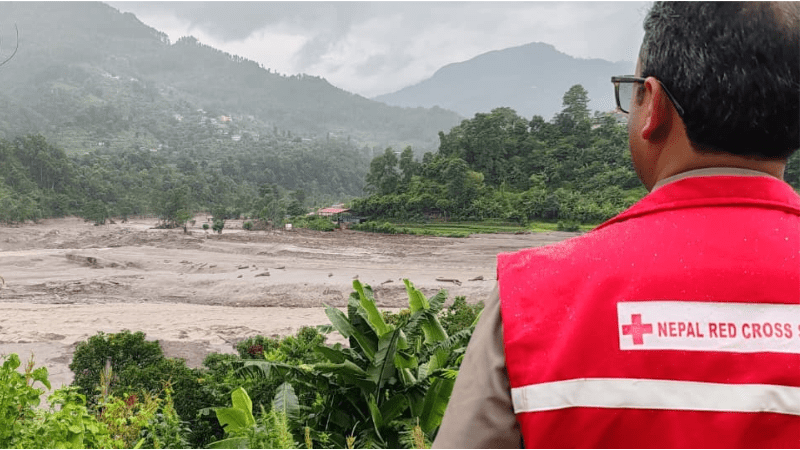 Person in a red Nepal Red Cross vest observing a wide river in a mountainous landscape following flooding.