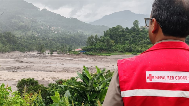 Person in a red Nepal Red Cross vest observing a wide river in a mountainous landscape following flooding.