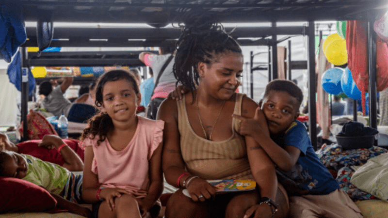 Mother and two children sitting together inside a temporary shelter, with beds and personal belongings visible in the background.