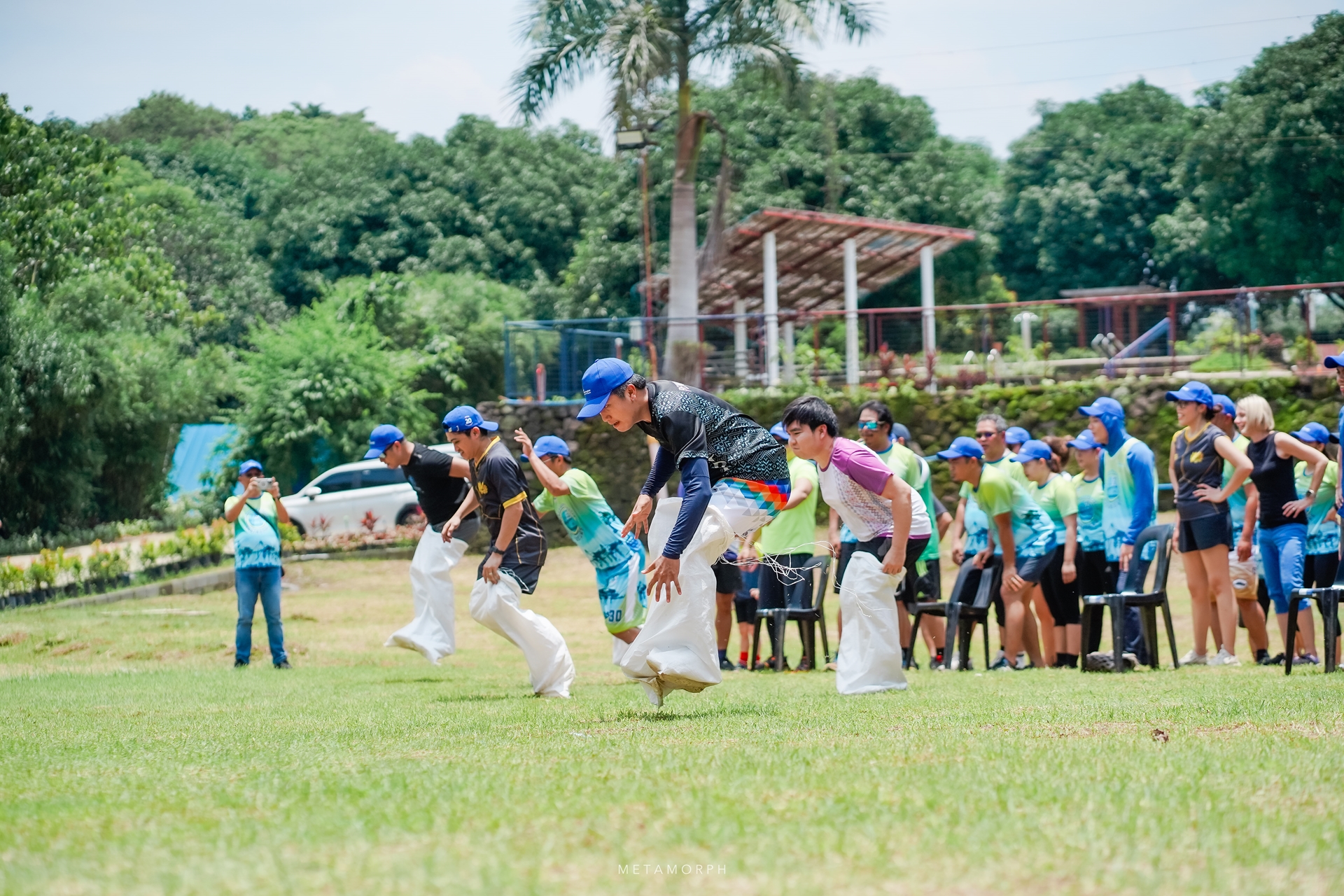 everal people wearing blue baseball caps are actively jumping forward on a grassy field, each enclosed in a white burlap sack up to their waist. 