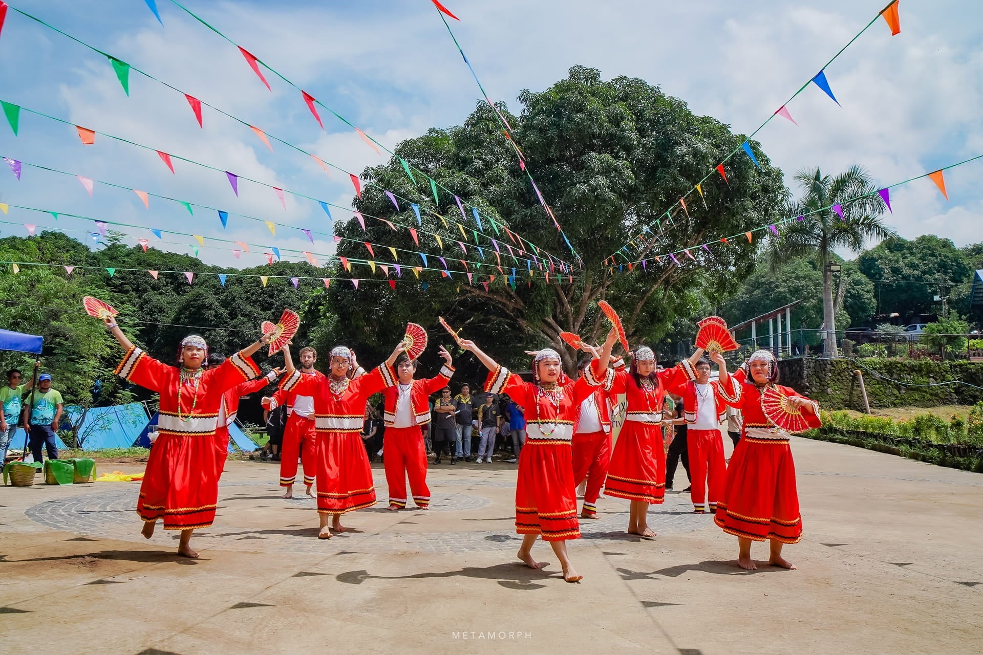 A group of eight performers in vibrant red and white traditional clothing are dancing outdoors, holding decorative fans high above their heads.