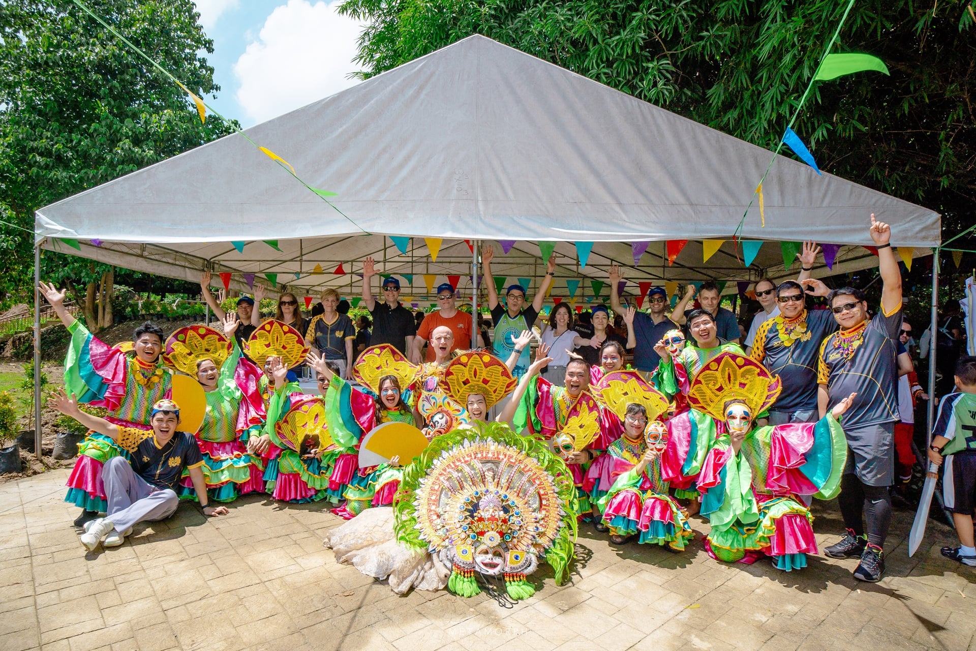 Eleven costumed dancers with bright pink/green outfits and headdresses pose with a large group of people under a white tent.