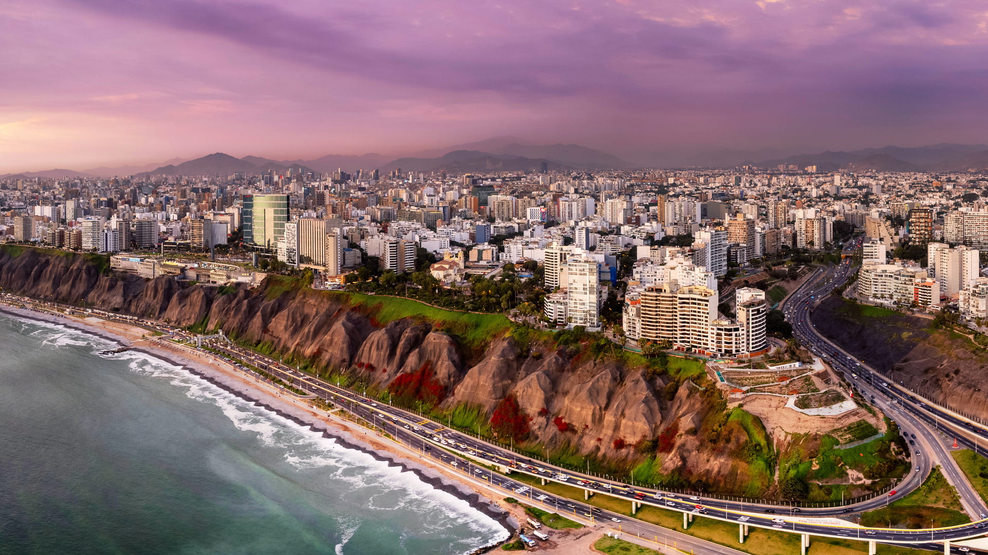 Panoramic aerial view of Lima, Peru, showing city buildings, the coastal cliffs, and a highway along the Pacific Ocean.