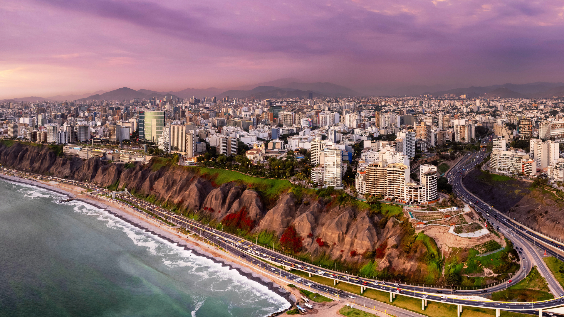 Panoramic aerial view of Lima, Peru, showing city buildings, the coastal cliffs, and a highway along the Pacific Ocean.