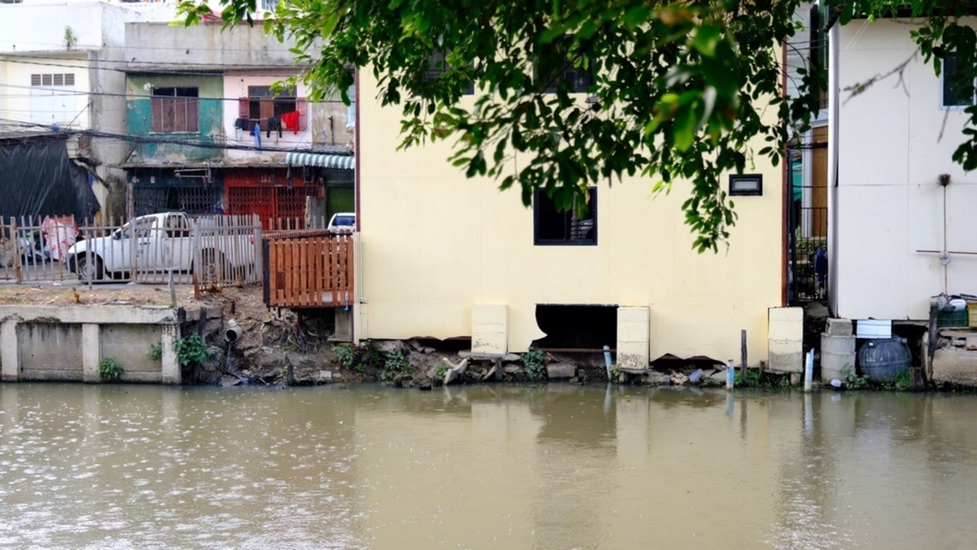 Flood‑prone riverside housing along an urban canal in Thailand, showing erosion, weakened building foundations, and the urgent need for resilient flood management.
