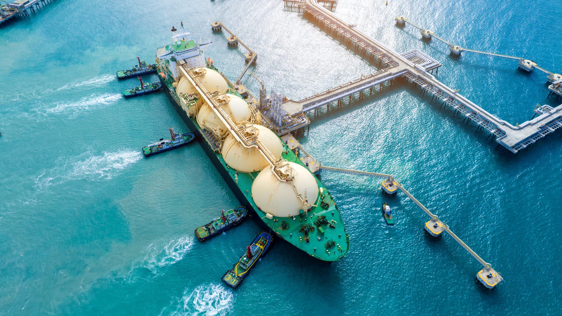 Aerial view of a large green liquefied natural gas (LNG) tanker ship docked at a port terminal. The ship has four large spherical tanks on its deck. Several tugboats are assisting the tanker in maneuvering into position at the dock. The water around the ship is a vibrant blue, and the port infrastructure includes multiple piers and docking platforms extending into the water.