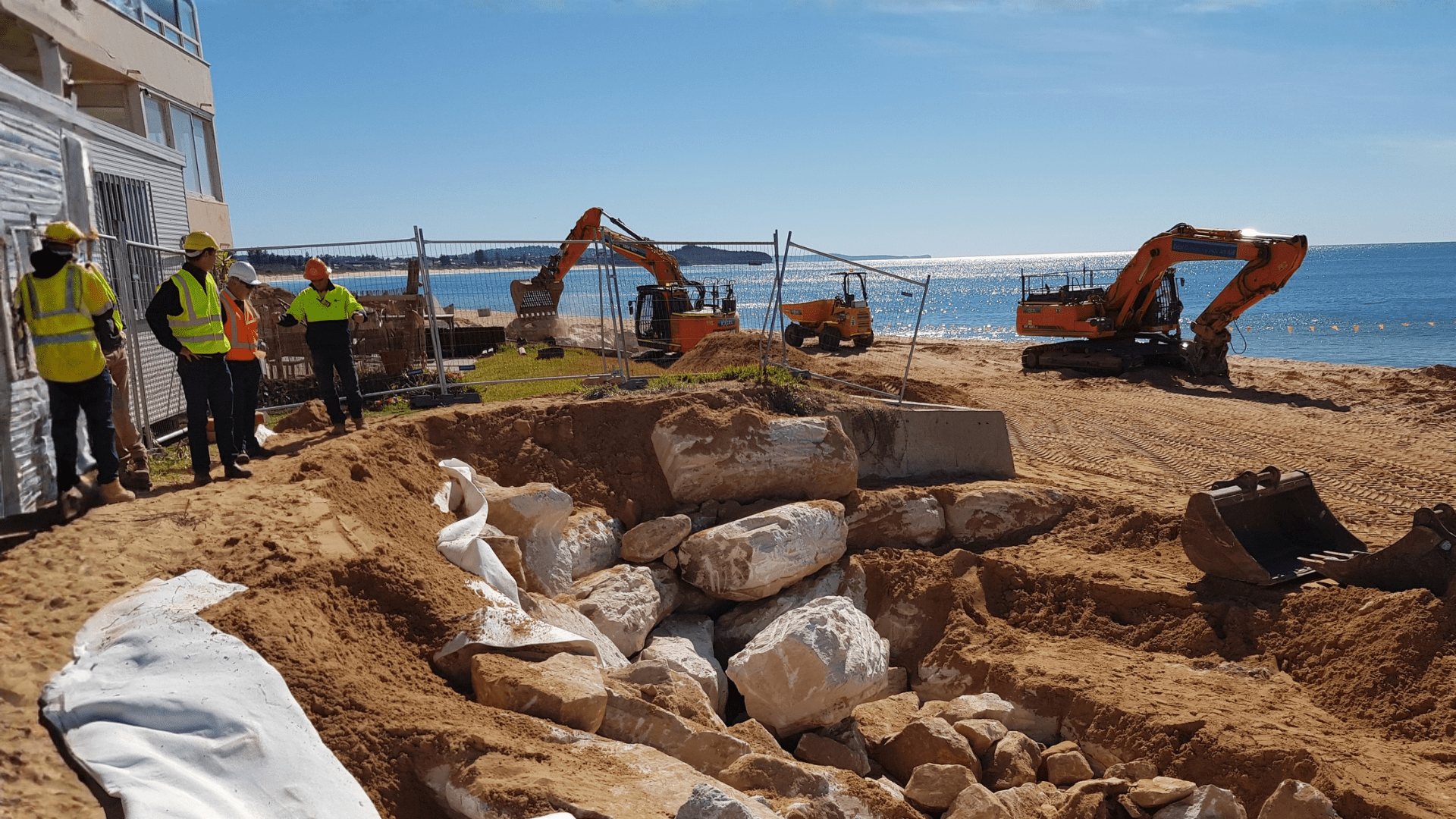 Coastal construction works on a sandy beach, with excavators and workers installing a rock revetment near the shoreline to support erosion and coastal protection measures.