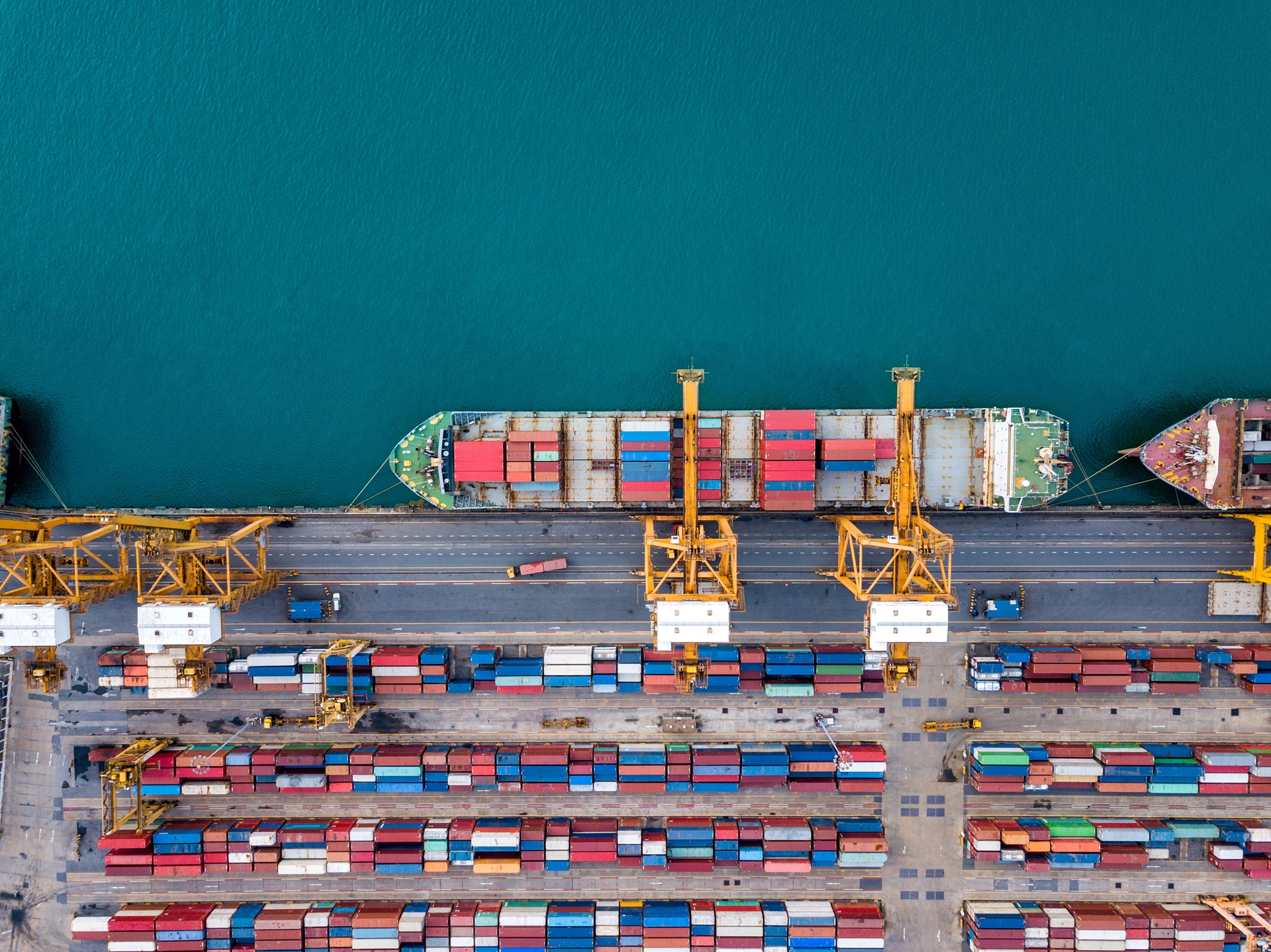Aerial view of a container ship docked at a port being loaded by large cranes, with rows of colourful shipping containers stacked along the quay.