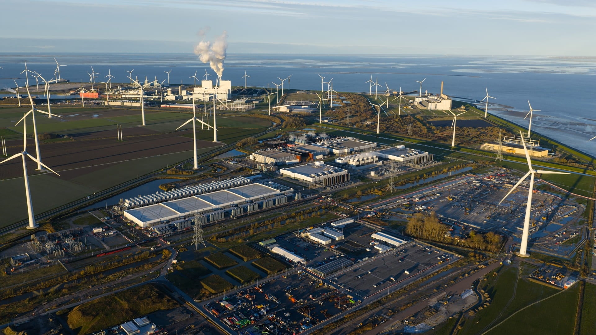 Aerial view of a large-scale European data centre campus integrated within an energy infrastructure landscape with wind turbines and industrial facilities, illustrating sustainable digital infrastructure expansion.