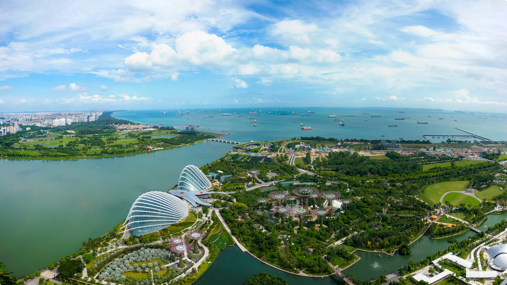 Aerial view of Singapore coastline featuring Gardens by the Bay, Marina Barrage, and surrounding green spaces with ships offshore in the Singapore Strait.