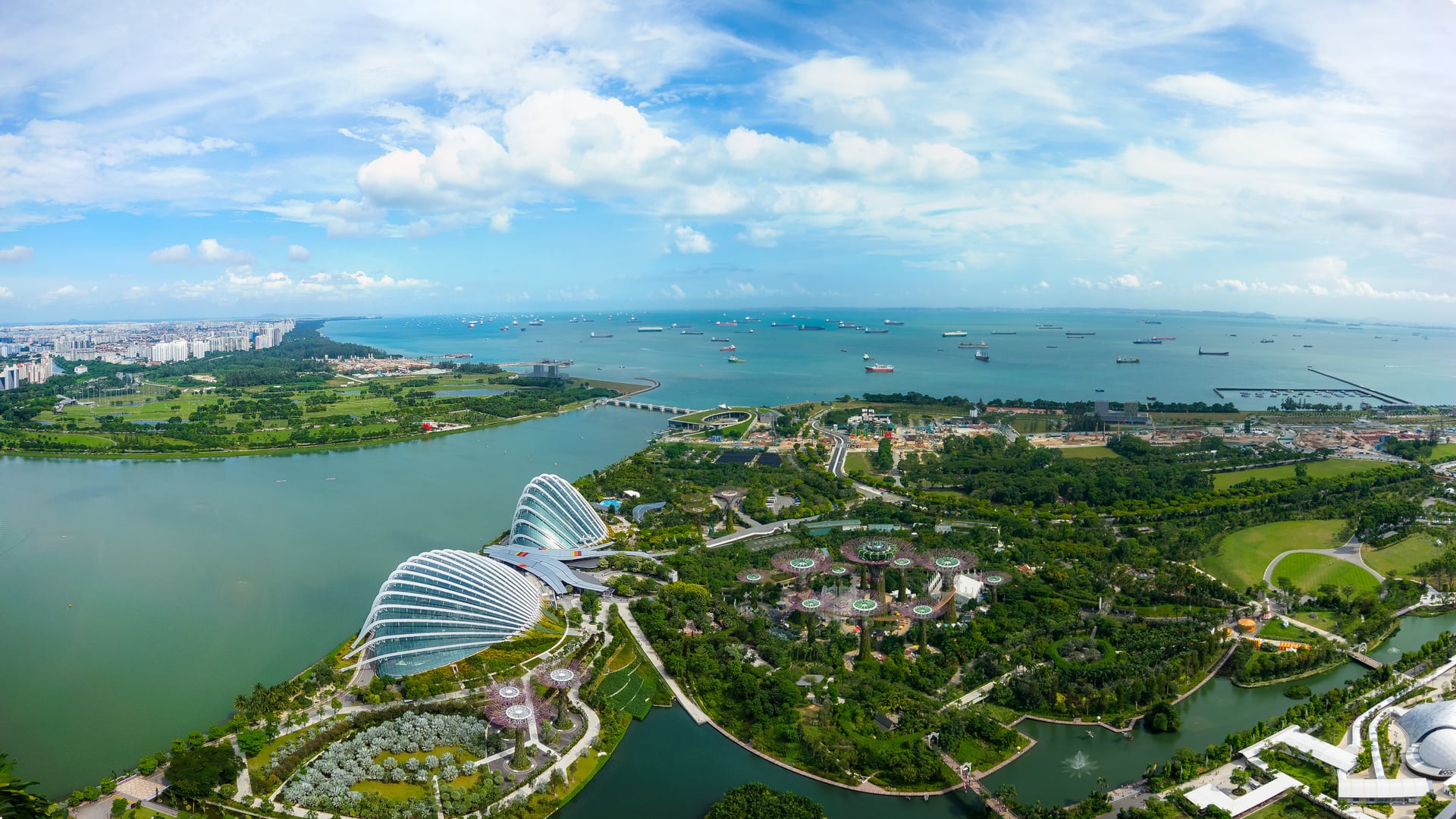 Aerial view of Singapore coastline featuring Gardens by the Bay, Marina Barrage, and surrounding green spaces with ships offshore in the Singapore Strait.