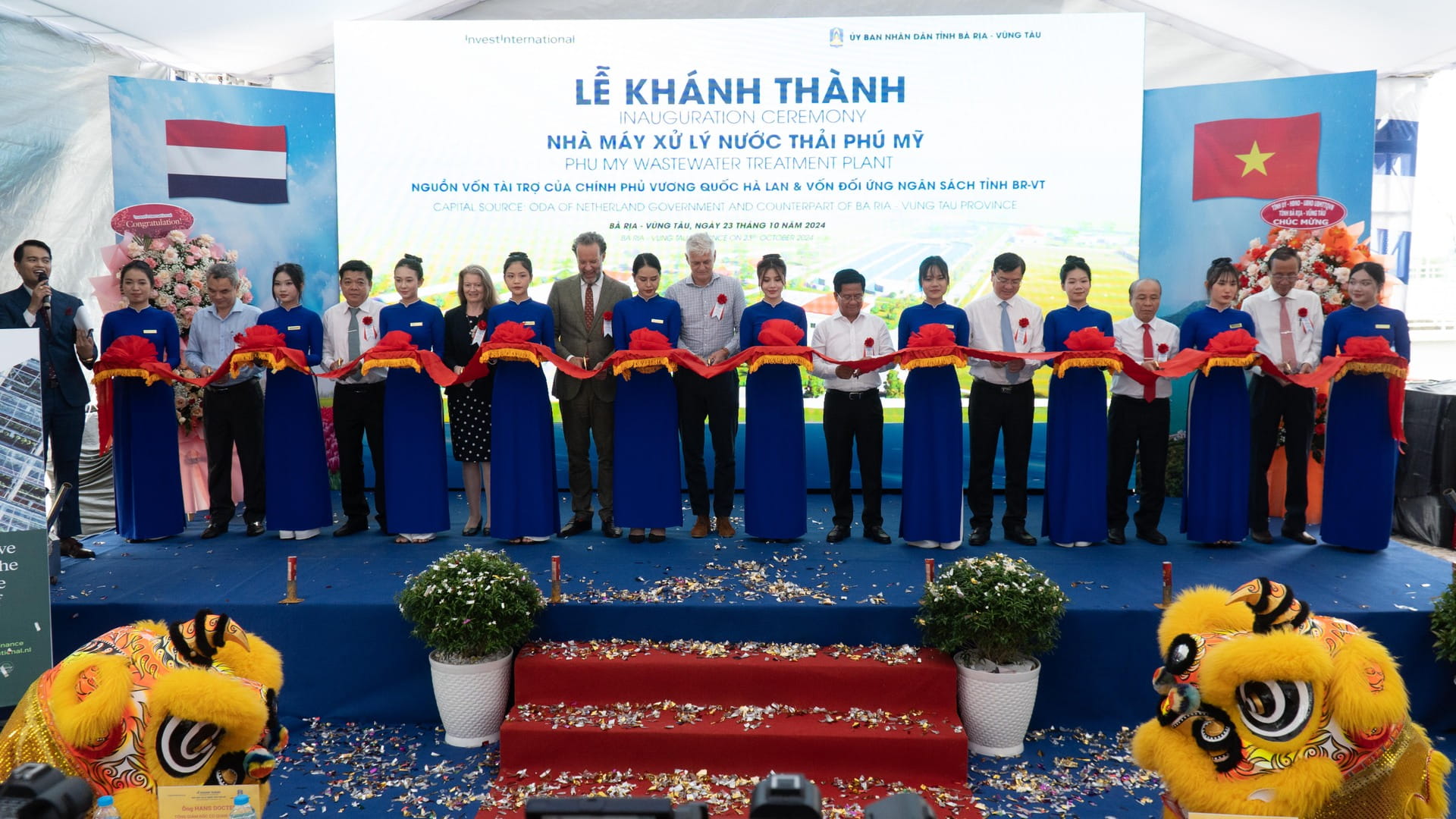 A group of people in formal attire stands on stage cutting a ribbon at an inauguration ceremony with Vietnamese flags and two lion dance costumes in front.