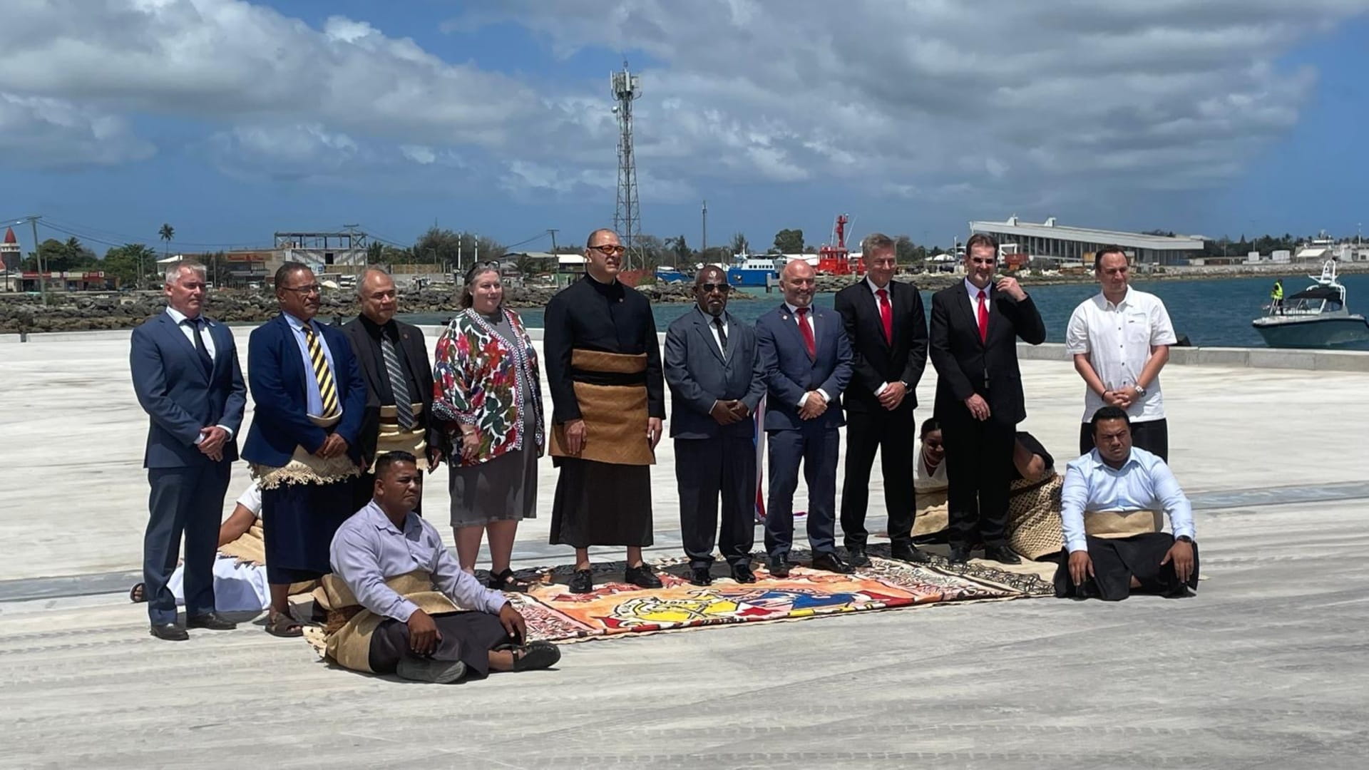 Group of officials and community members standing and sitting on a newly upgraded port dock in Tonga, with traditional woven mats and attire displayed. The background shows a harbor with boats, industrial buildings, and a communication tower under a partly cloudy sky, highlighting maritime infrastructure improvements