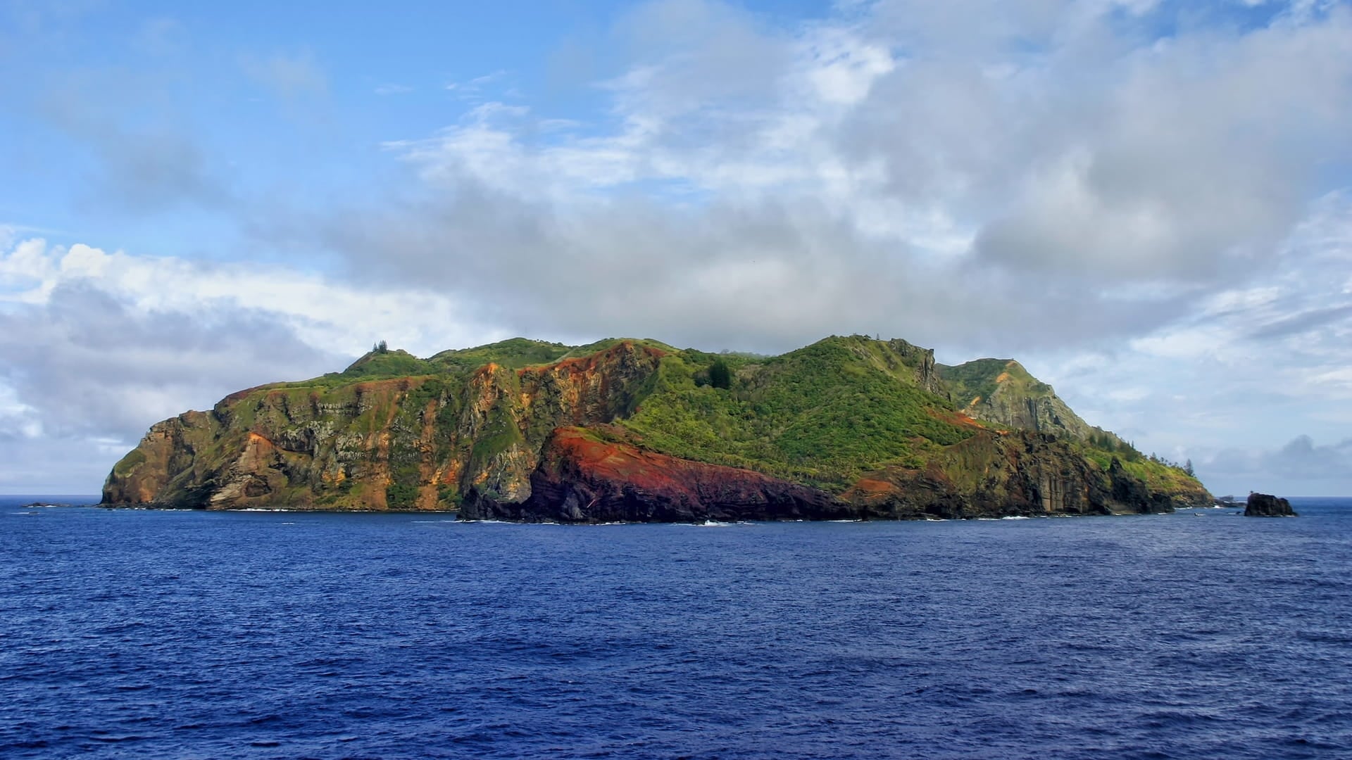 Pitcairn Islands with green hills and exposed red and brown rock cliffs, surrounded by the open Pacific Ocean beneath a cloudy blue sky