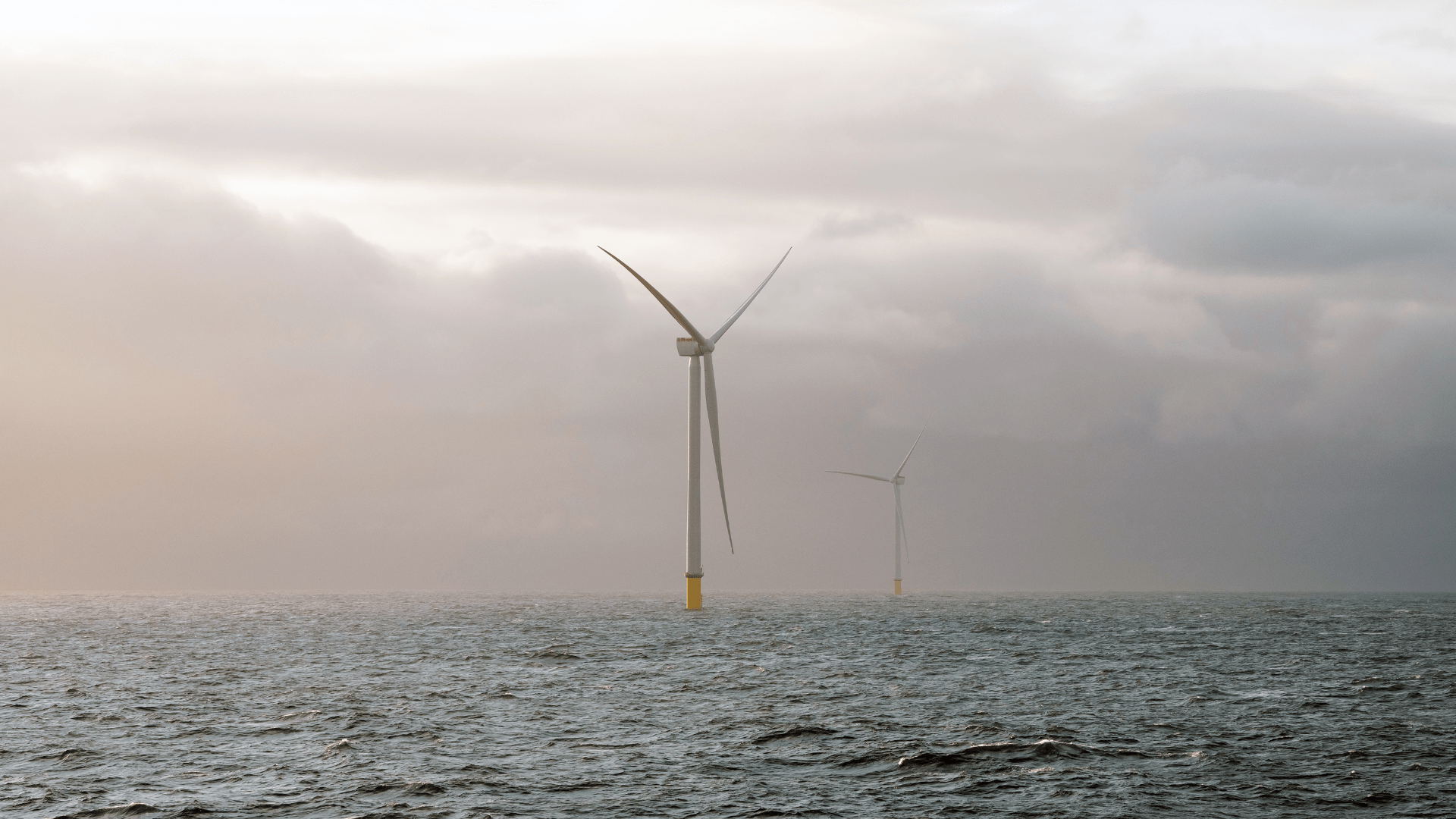 Two offshore wind turbines stand in the open sea, with calm water in the foreground and a cloudy, misty sky in the background.
