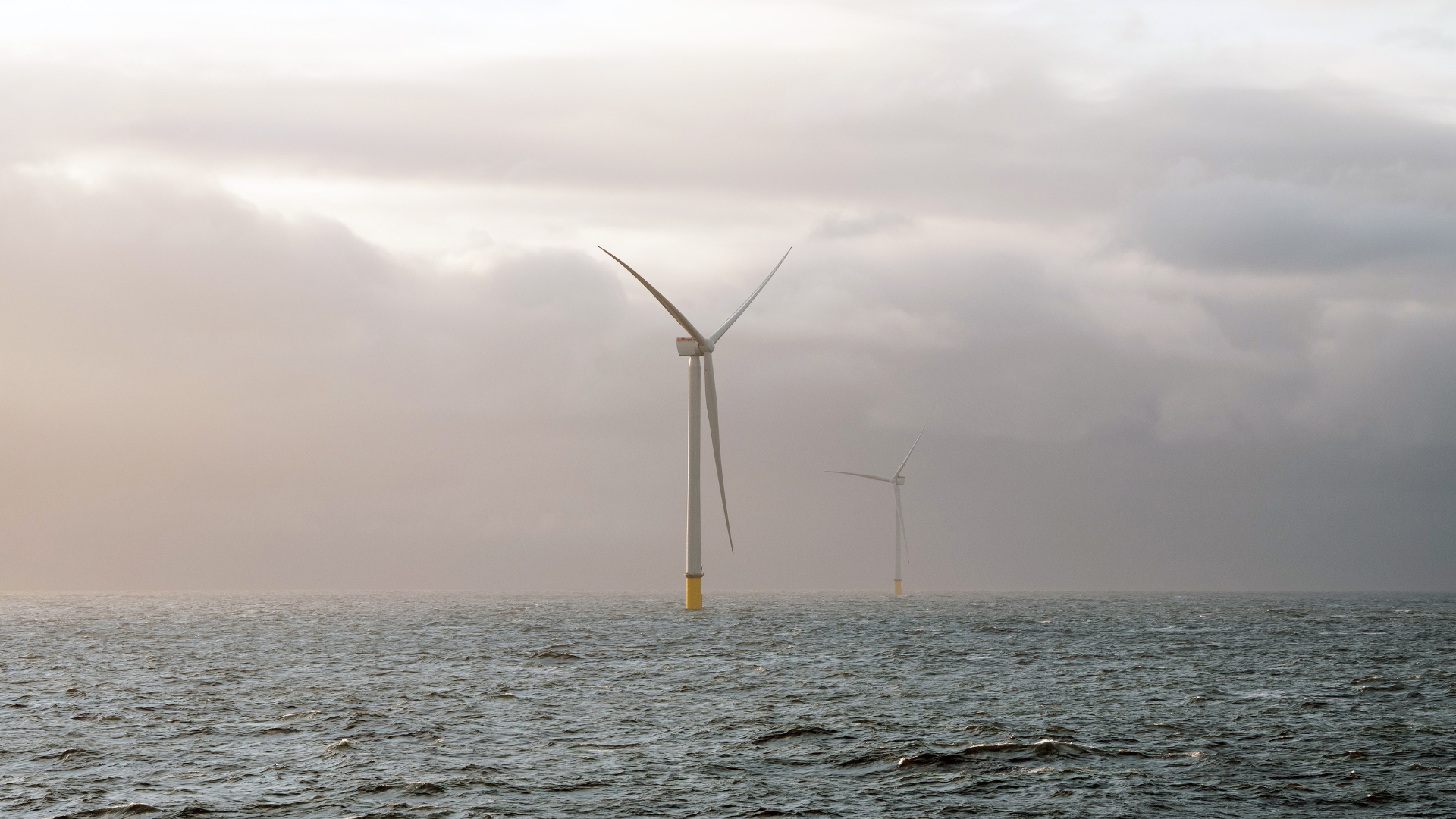 Two offshore wind turbines stand in the open sea, with calm water in the foreground and a cloudy, misty sky in the background.