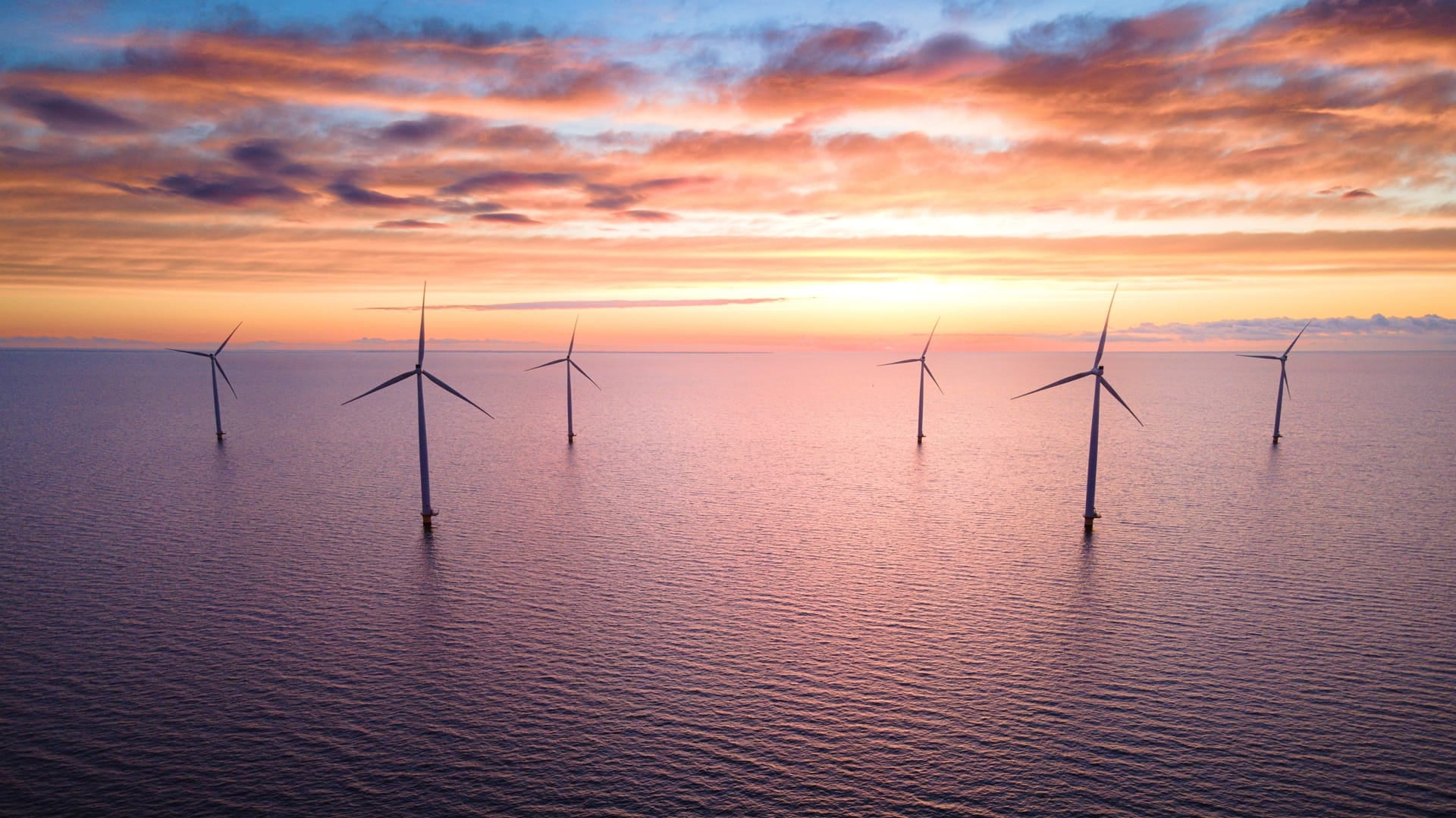 Aerial view of a line of offshore wind turbines in calm open water at sunset, with warm colours in the sky and reflections on the surface.