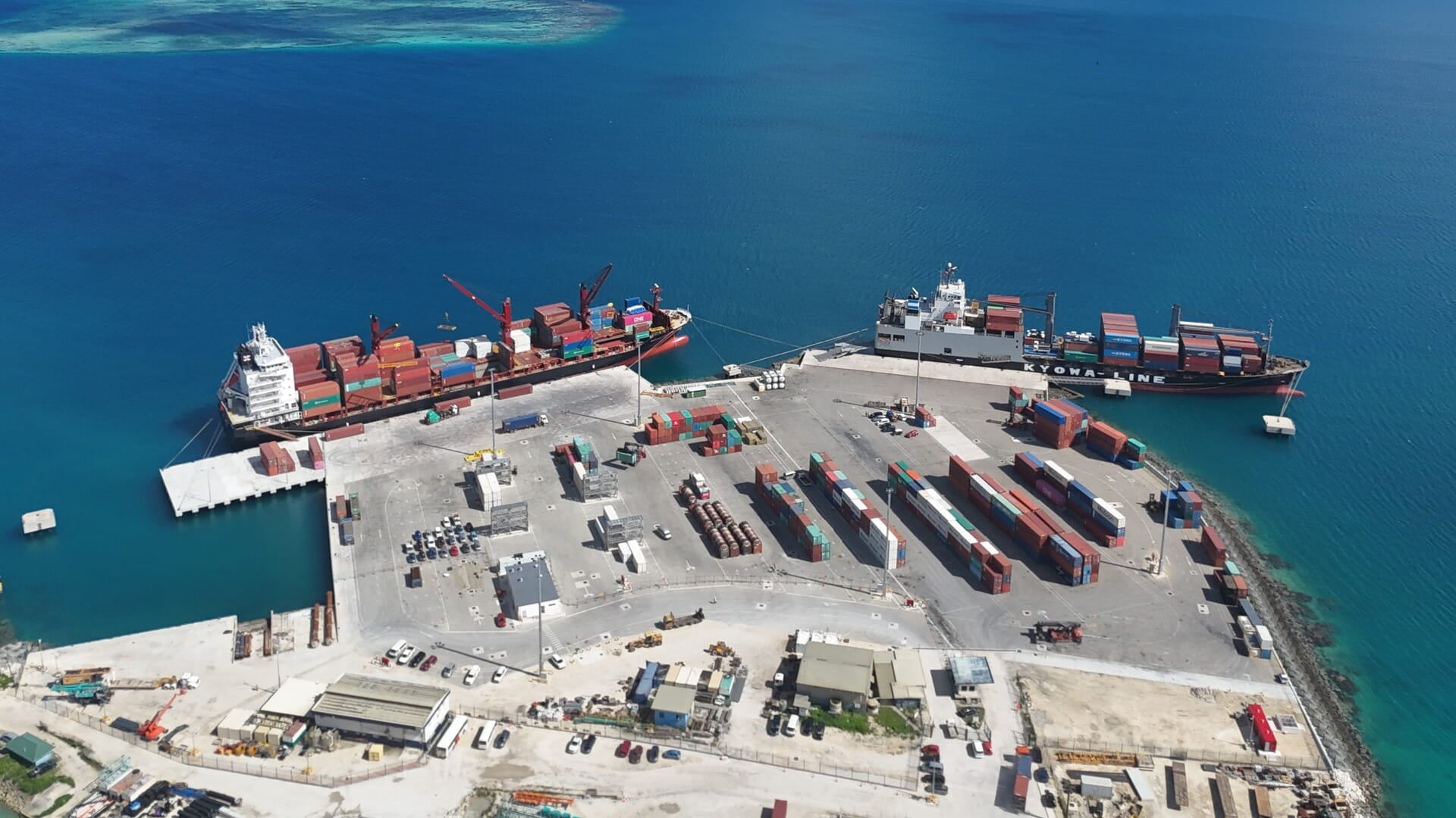 Aerial view of Tonga port showing two cargo ships docked at the pier, surrounded by stacked shipping containers and port facilities, with clear blue ocean in the background.