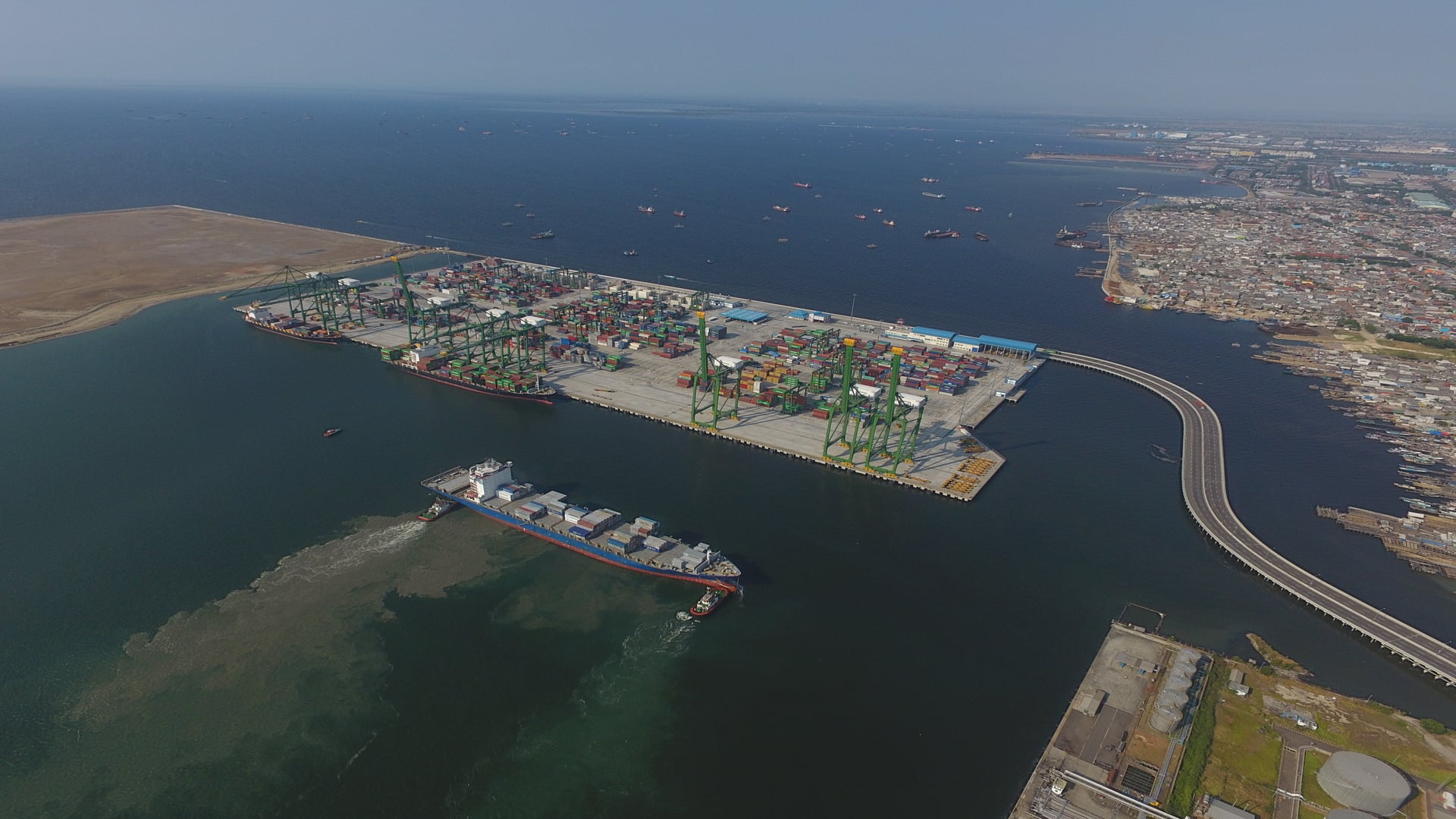 An aerial photograph of Kalibaru Phase 1A, featuring shipping containers, cranes, and a container ship near the terminal, with a curved access bridge and cityscape in the background.
