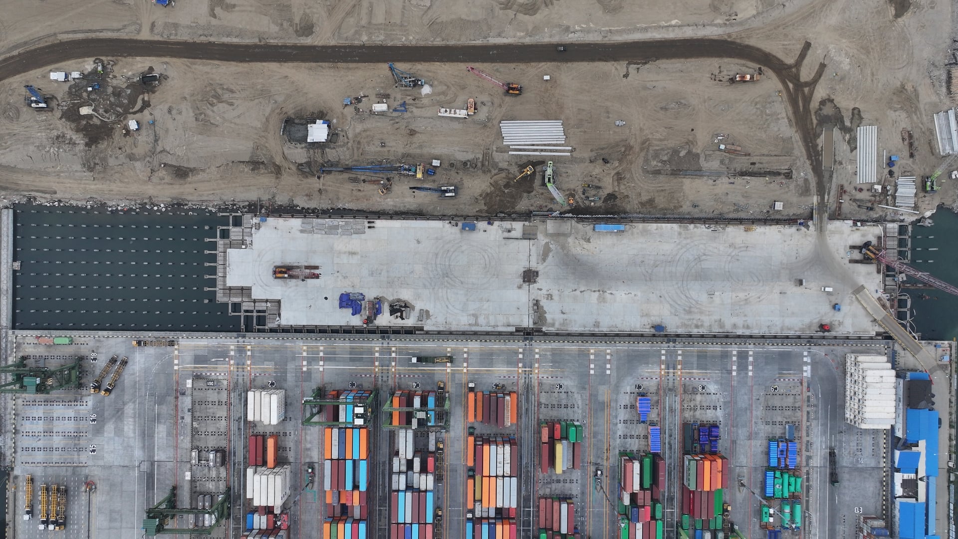 Aerial view of Kalibaru Phase 1B showing an active construction site with concrete areas, heavy machinery, and adjacent rows of colourful shipping containers at the operational section of the port.
