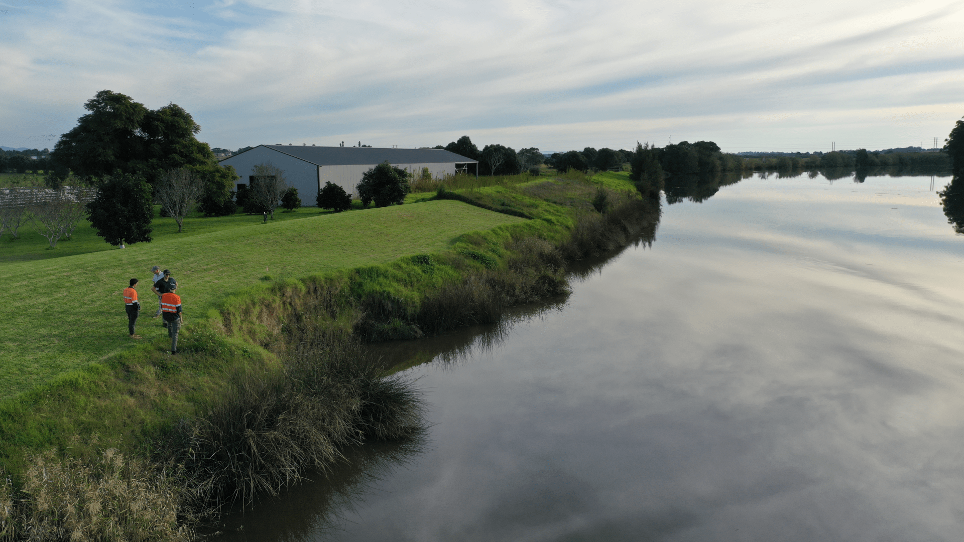Aerial view of a calm river bordered by grassy banks and rural land. Three field staff wearing high-visibility safety clothing stand near the river’s edge inspecting the embankment, while a large agricultural building and trees are visible in the background under a cloudy sky reflected in the water.