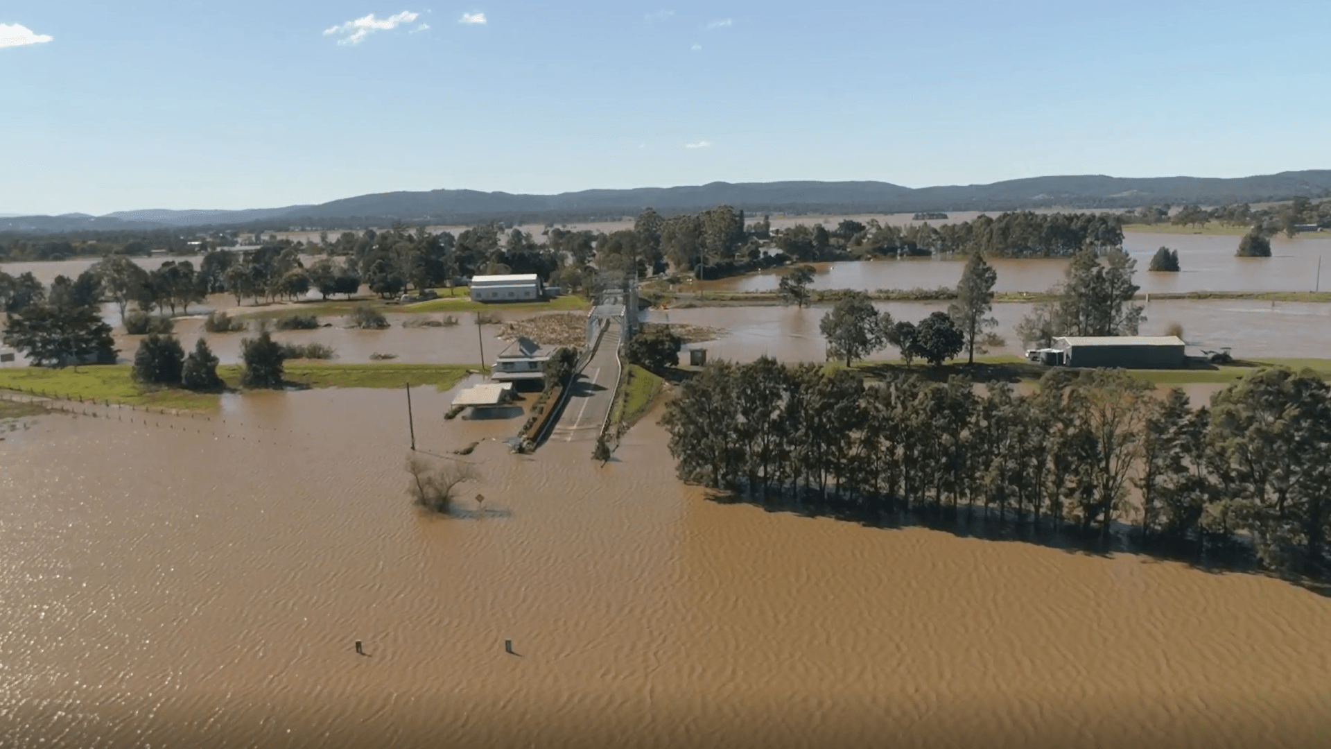 Aerial view of widespread flooding across a rural landscape, with muddy floodwaters surrounding fields, trees, farm buildings, and a road bridge crossing a swollen river. Low hills are visible in the distance under a clear blue sky.