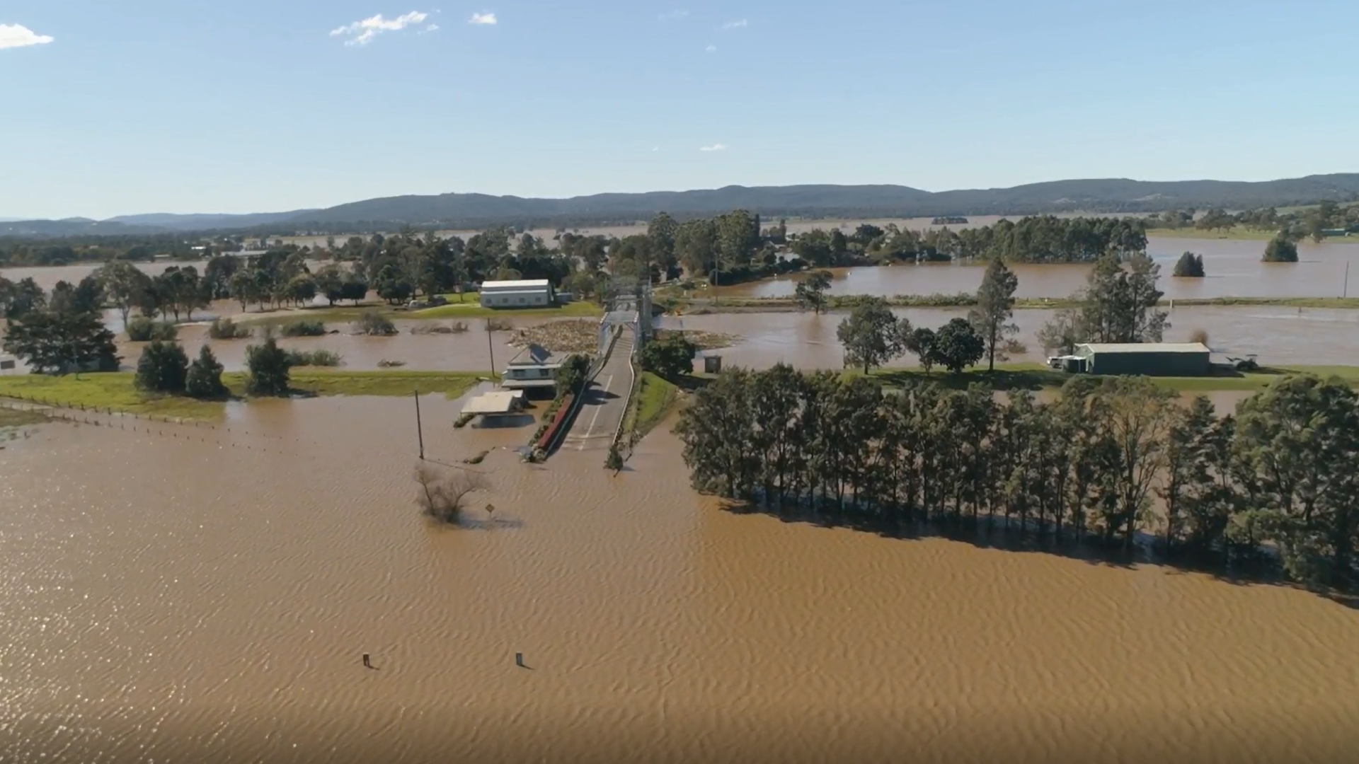 Aerial view of widespread flooding across a rural landscape, with muddy floodwaters surrounding fields, trees, farm buildings, and a road bridge crossing a swollen river. Low hills are visible in the distance under a clear blue sky.