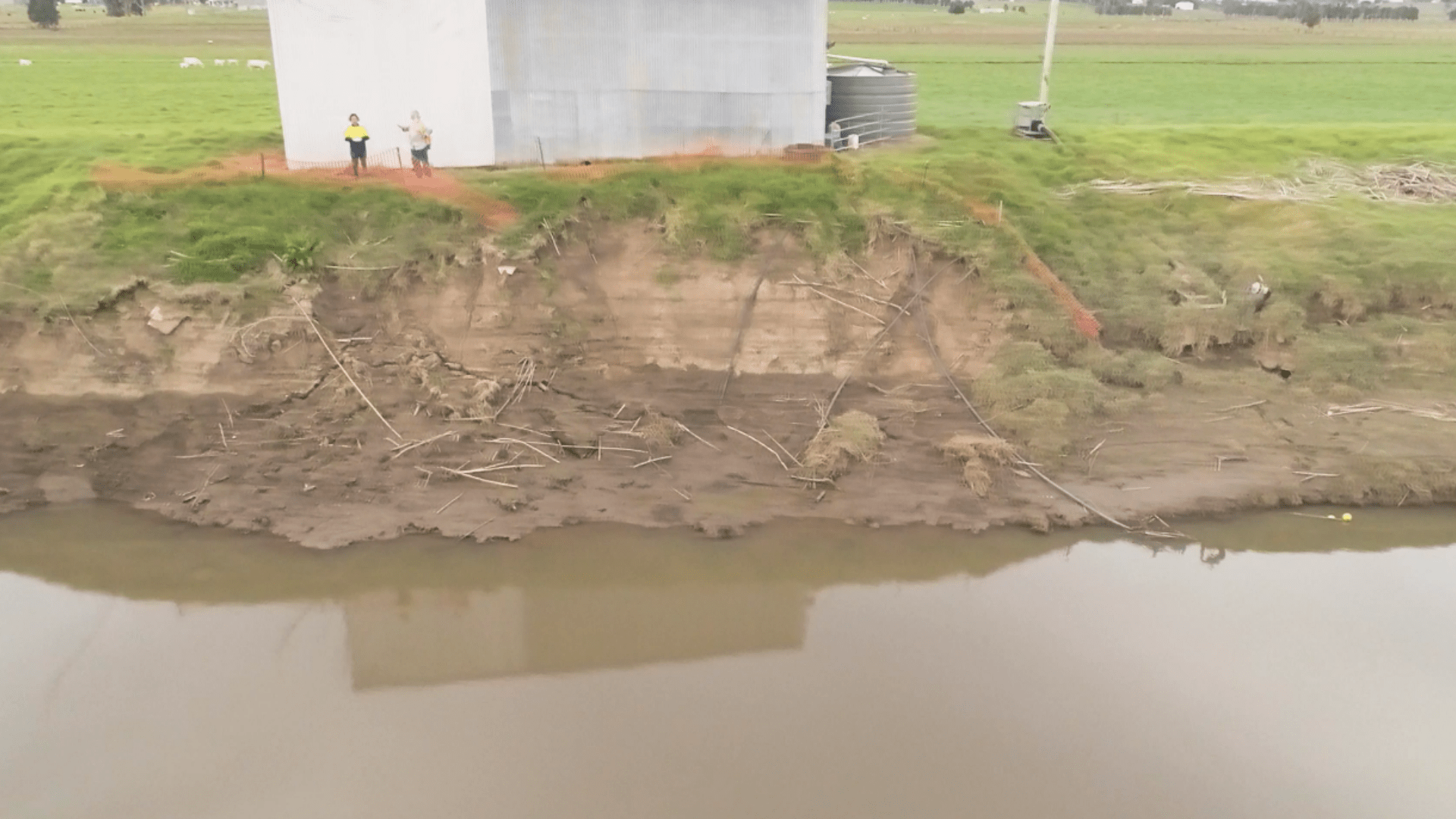 Severe riverbank erosion along a muddy waterway beside a rural property. A large section of the embankment has collapsed, exposing soil layers beneath grassy ground near storage tanks, while two people stand at the edge observing the damage.