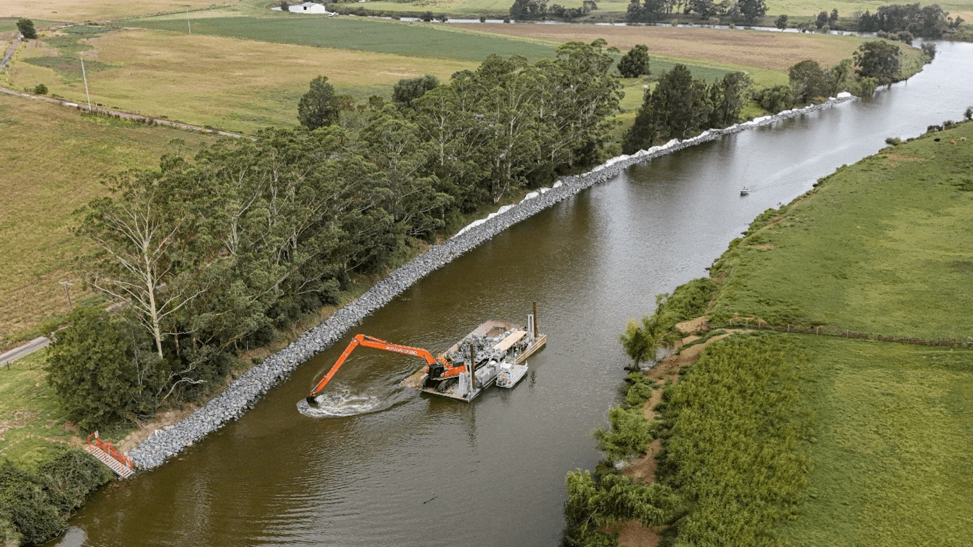Aerial view of a riverbank stabilization project in a rural area, with rock armoring (riprap) lining the shore and an excavator mounted on a floating barge working in the water. Agricultural fields and rows of trees border the river on both sides.