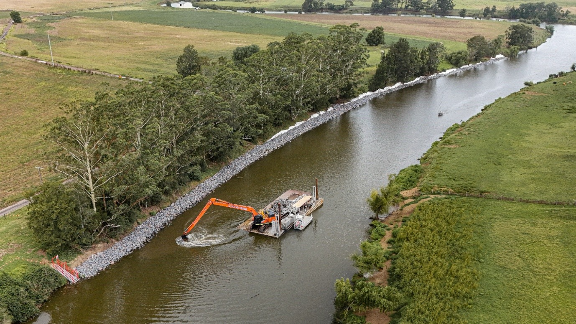 Aerial view of a riverbank stabilization project in a rural area, with rock armoring (riprap) lining the shore and an excavator mounted on a floating barge working in the water. Agricultural fields and rows of trees border the river on both sides.