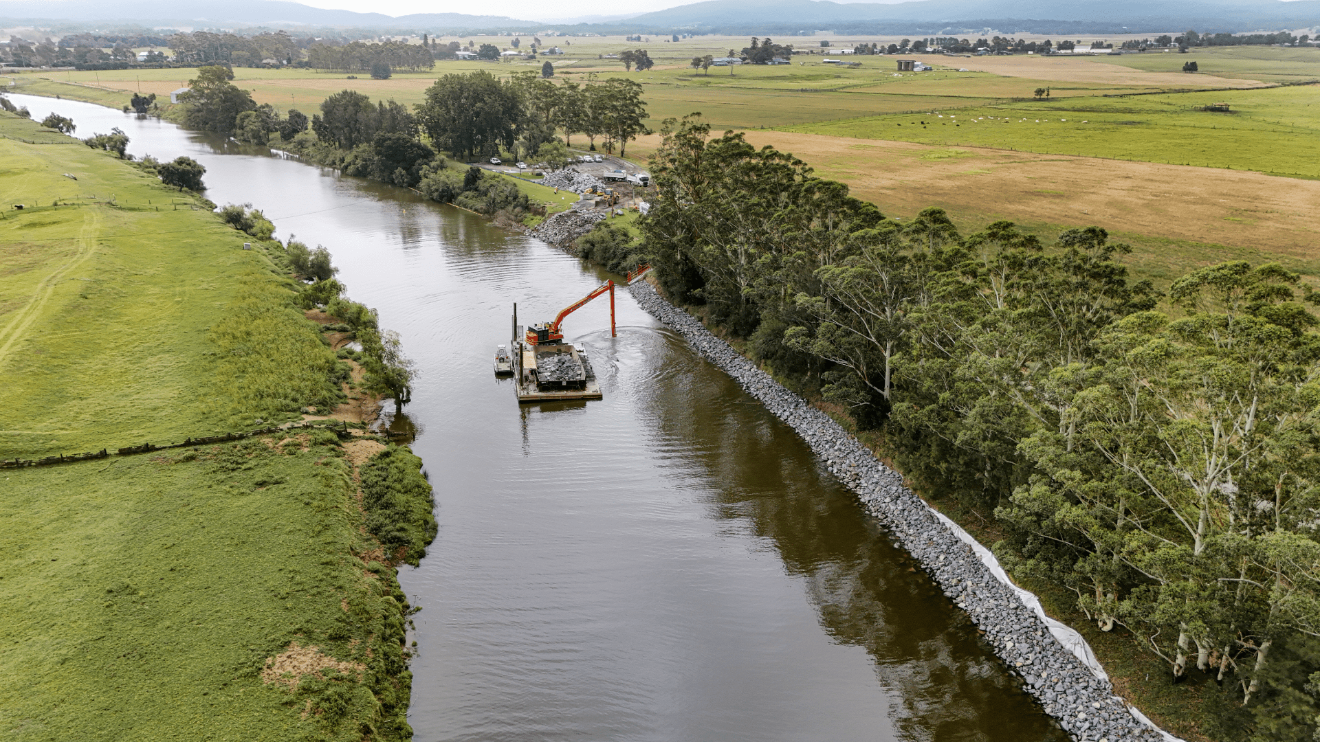 Aerial view of a riverbank stabilization project in a rural landscape. An excavator mounted on a floating barge is placing rock riprap along the right riverbank, while completed rock armoring extends beside a line of mature trees. Surrounding the river are open agricultural fields, scattered farm buildings, and distant rolling hills under an overcast sky.