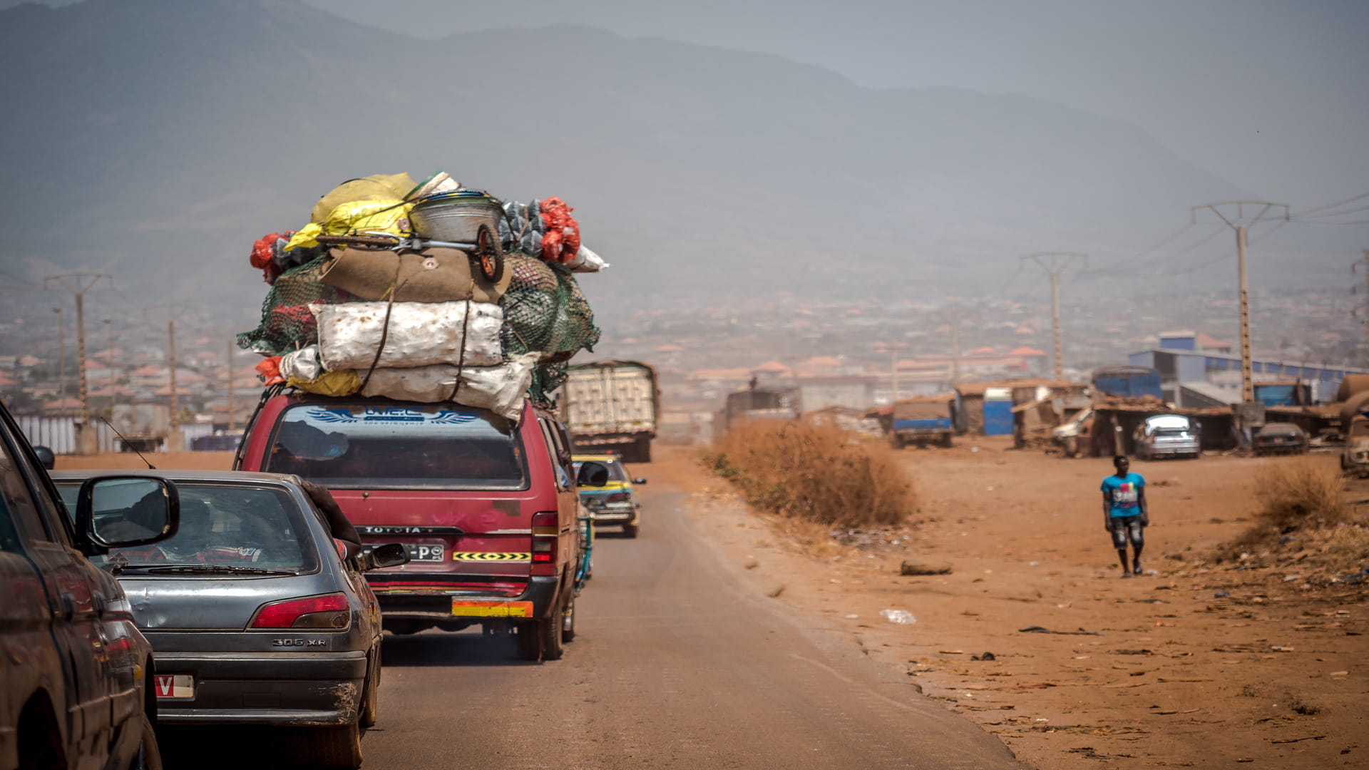 Overloaded minibus travelling along a rural road in Zambia, carrying goods on the roof with surrounding traffic and dry roadside landscape.