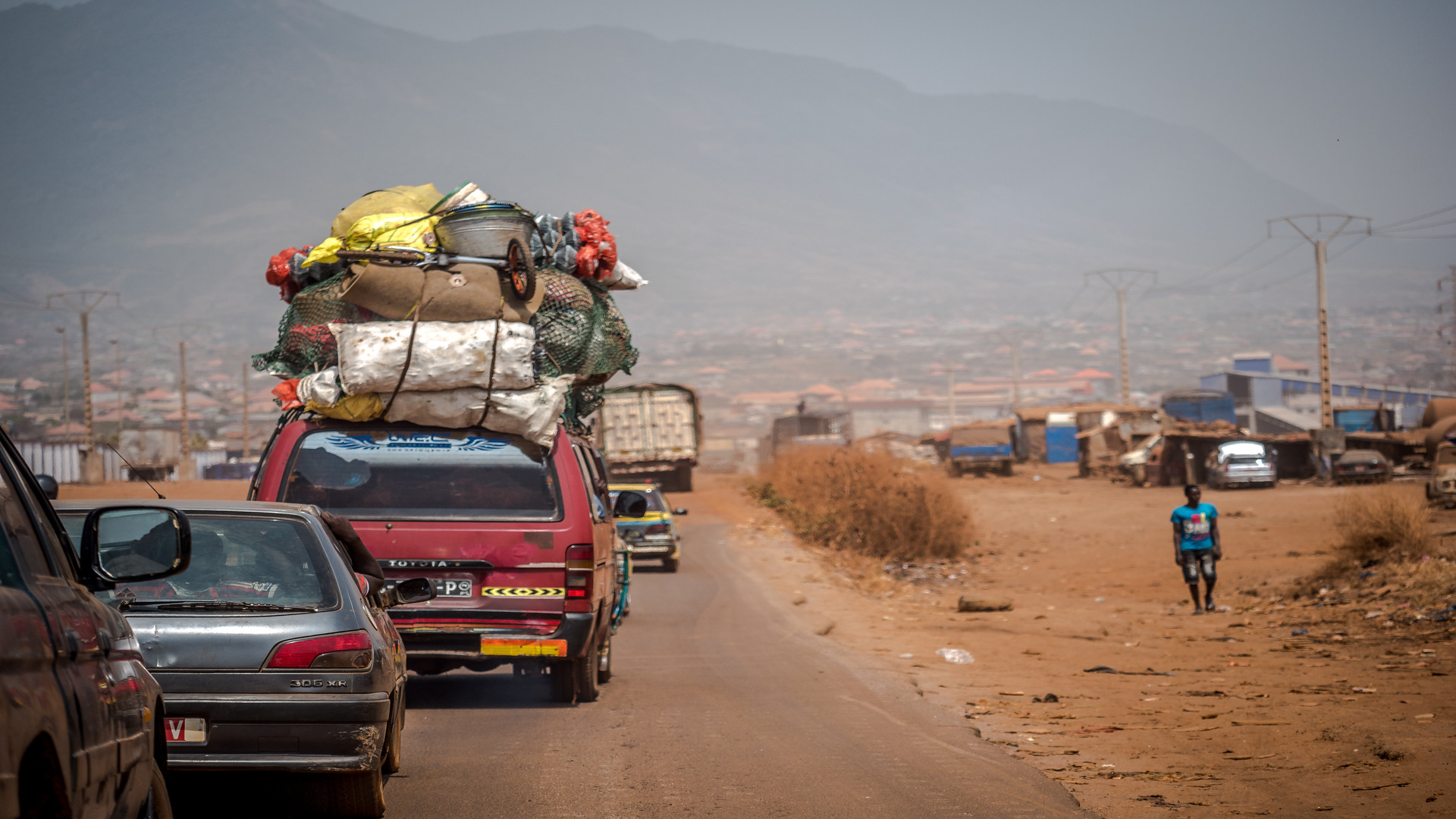 Overloaded minibus travelling along a rural road in Zambia, carrying goods on the roof with surrounding traffic and dry roadside landscape.