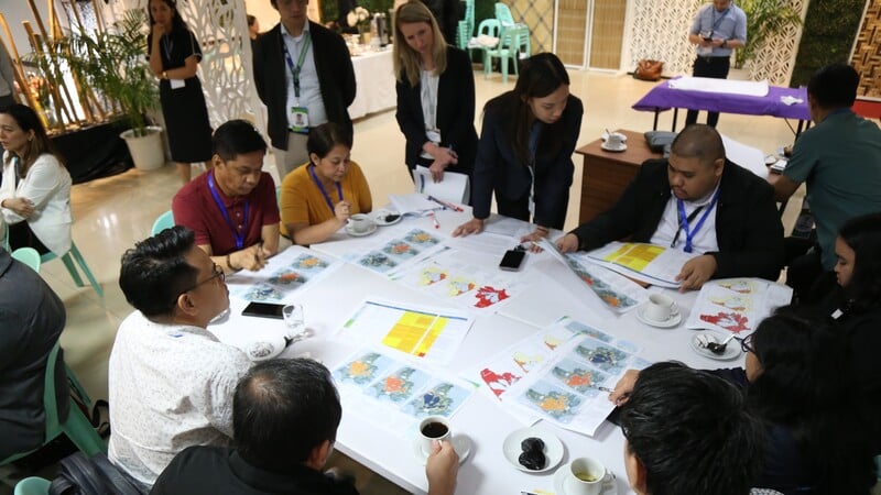 A group of people seated around a table in an indoor setting, engaged in a meeting or discussion. The table is covered with documents, some featuring colourful charts. Participants are taking notes, and cups of coffee and other beverages are visible. Plants and decorative elements are in the background.