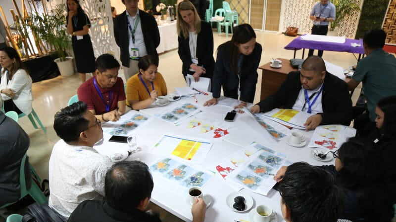 A group of people seated around a table in an indoor setting, engaged in a meeting or discussion. The table is covered with documents, some featuring colourful charts. Participants are taking notes, and cups of coffee and other beverages are visible. Plants and decorative elements are in the background.