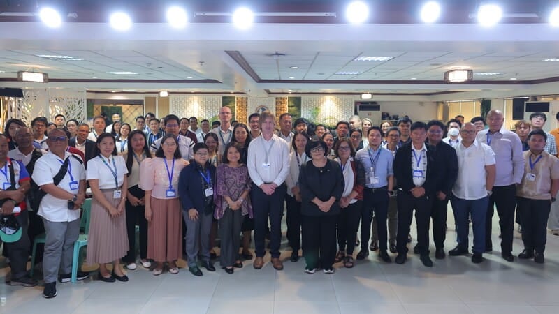 A large group of people in business casual attire standing together in a well-lit indoor venue. Most are wearing name tags or lanyards. The background includes a decorated wall with artwork and plants.