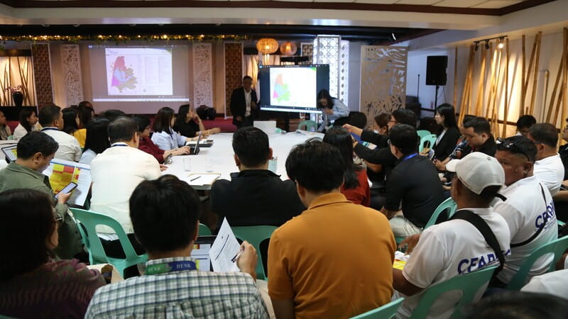 People seated around a large conference table in a meeting room, viewing two screens at the front that display maps and data. Attendees are actively engaged with documents and notes.
