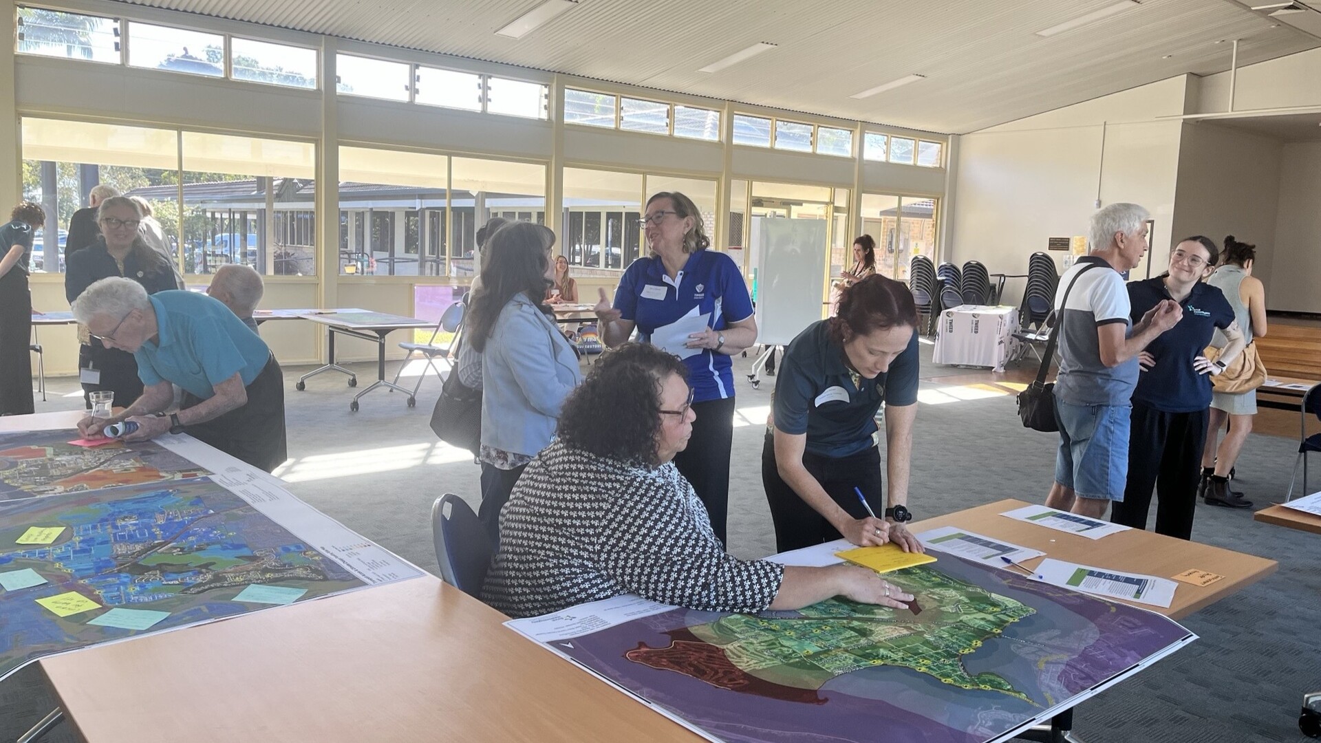 People gathered in a bright room, reviewing large maps and documents during a collaborative planning workshop, with discussions and note-taking around tables.