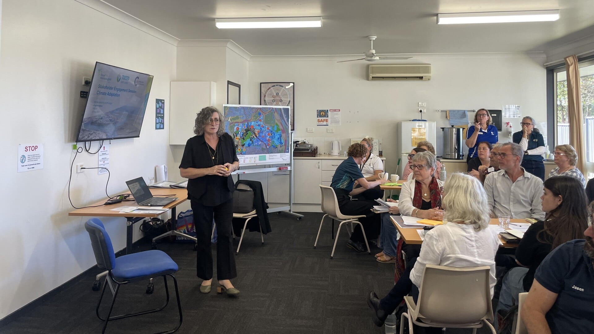 A presenter leads a climate adaptation workshop with a screen and map display, while attendees seated at tables listen, take notes, and engage in discussion.