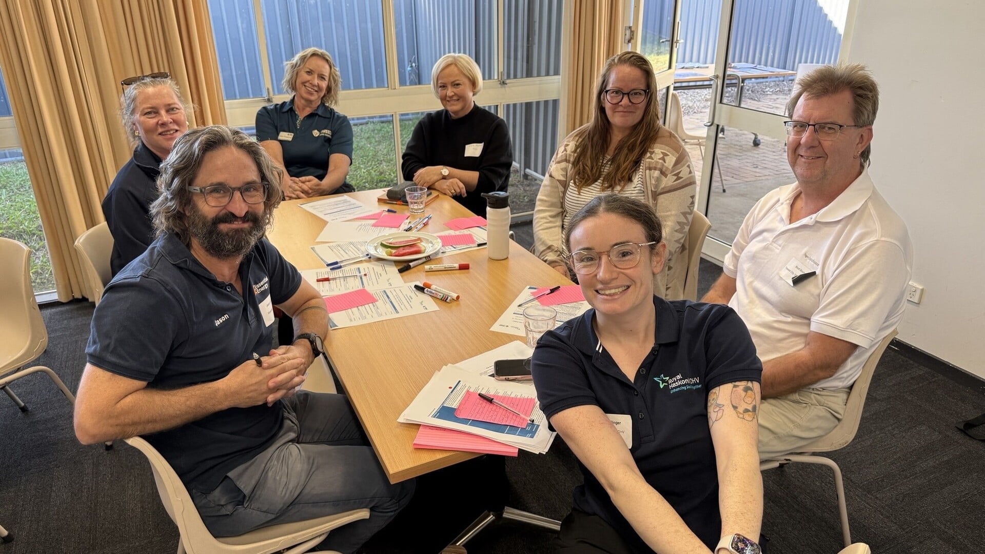 Seven people seated around a table with papers, notebooks, and snacks, engaged in a collaborative meeting or workshop in a room with large windows.