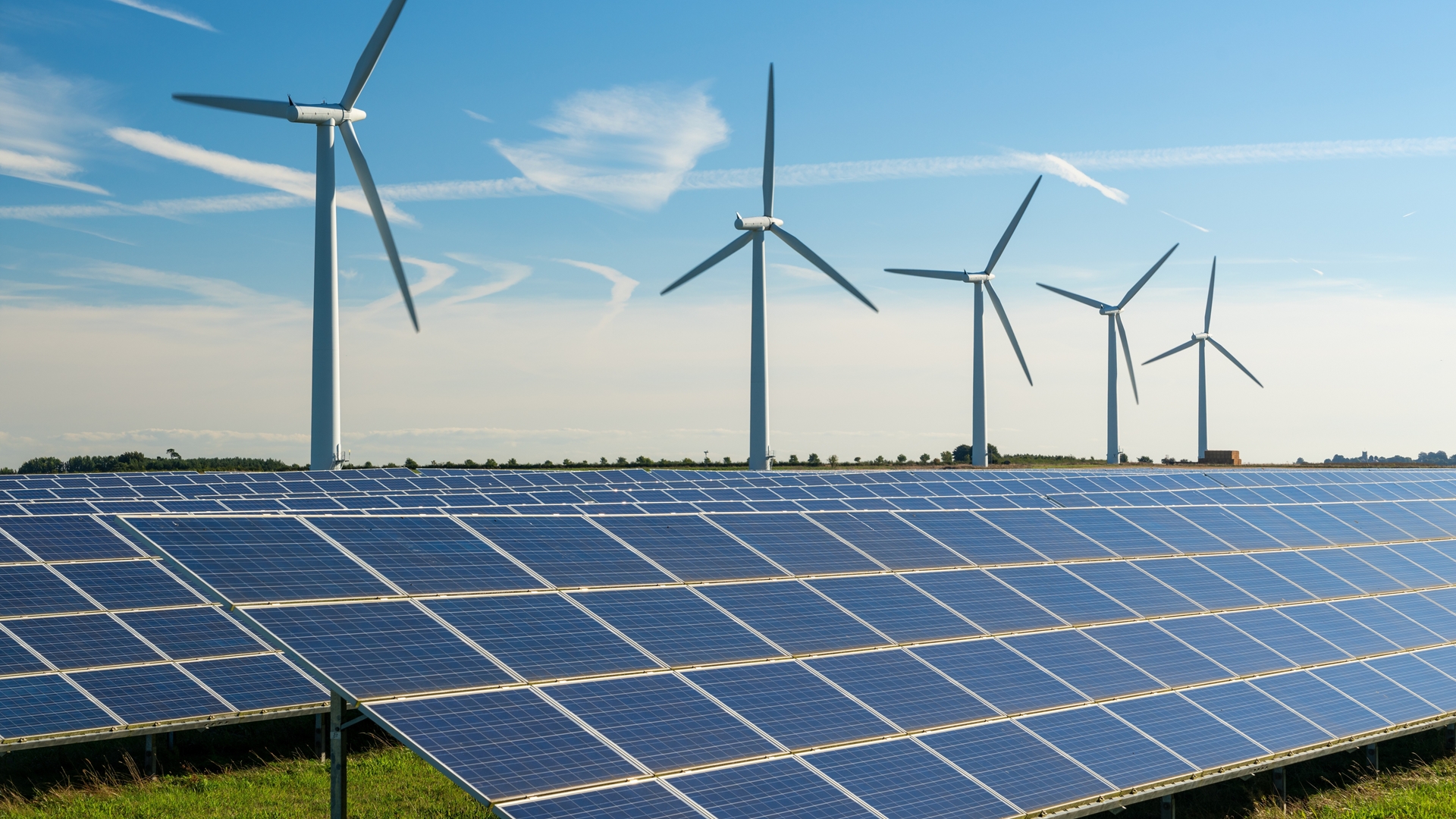 Solar panels in a field with several wind turbines in the background under a clear blue sky.