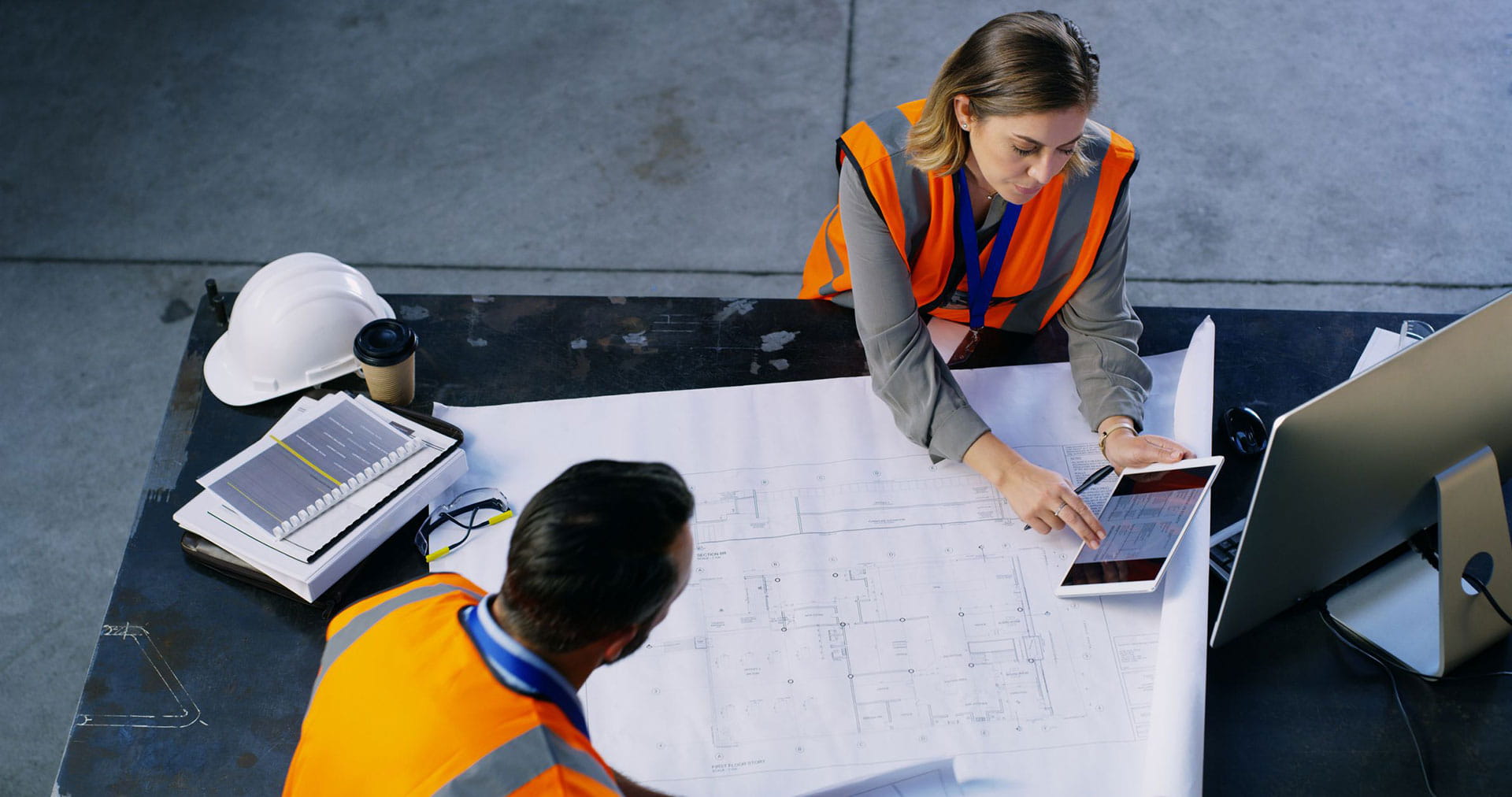 Two construction workers reviewing a blueprint at a desk.